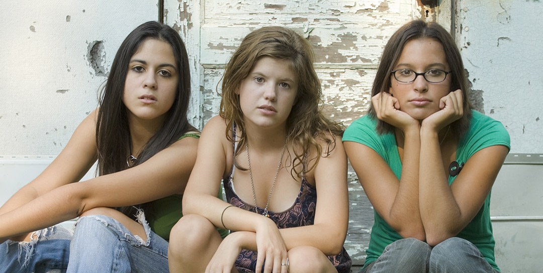three teen girls with serious facial expressions seated against grungy wall out of doors
